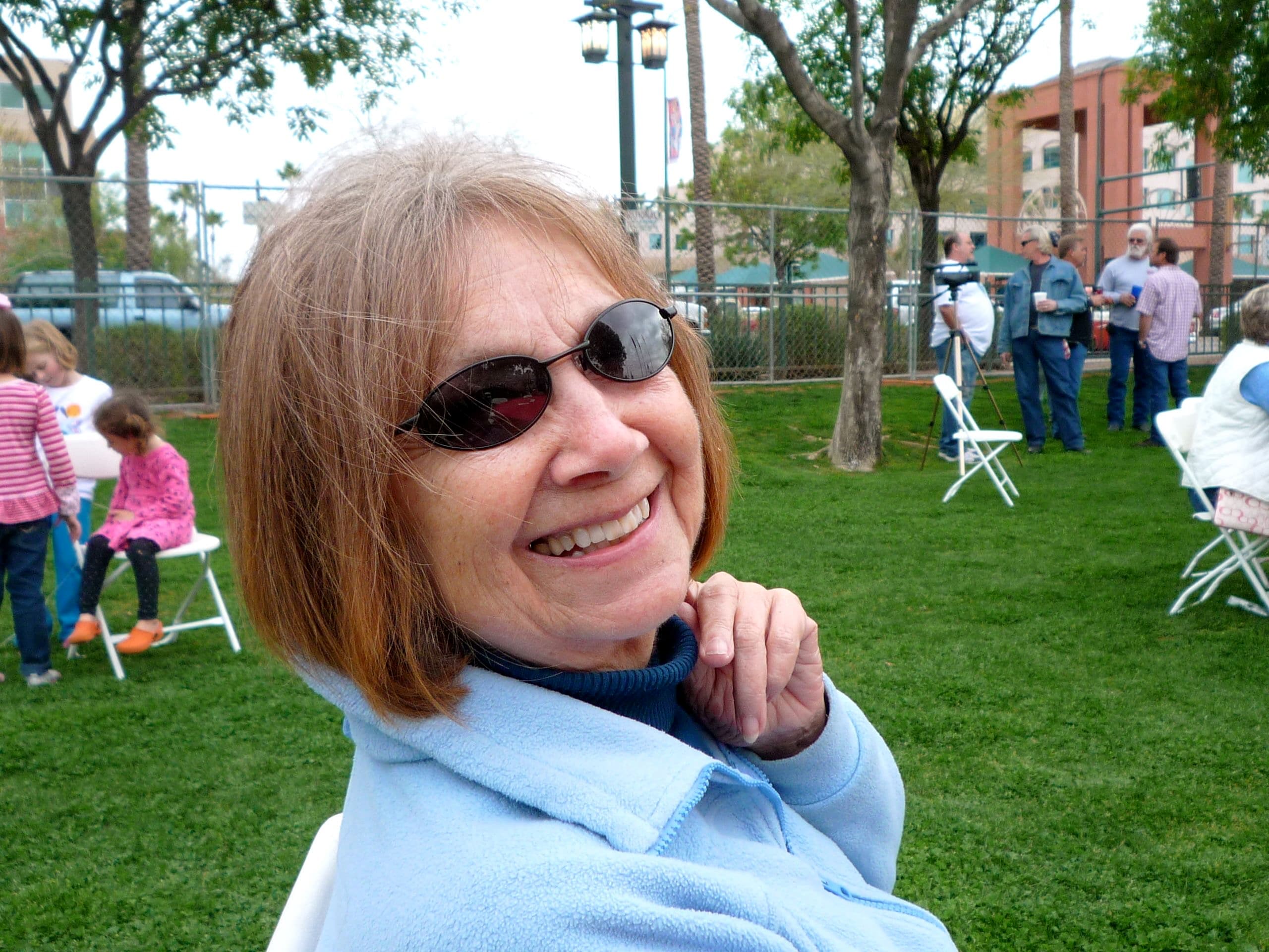 Wanda in sunglasses smiling over her shoulder at a park, children playing behind her